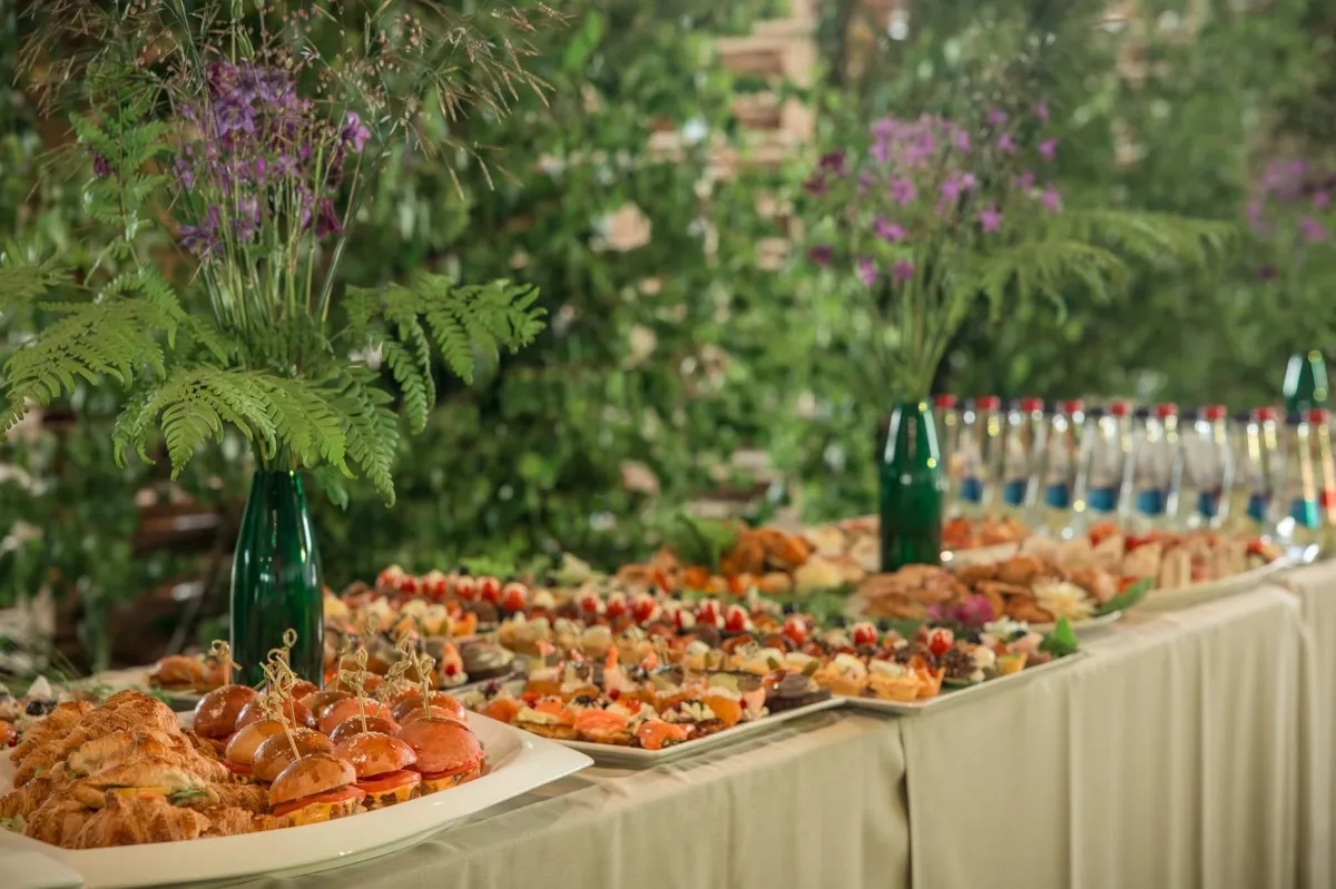 Bufetes galds ar mini burgeriem, kruasāniem un uzkodām, vāzēs pļavas ziedi
